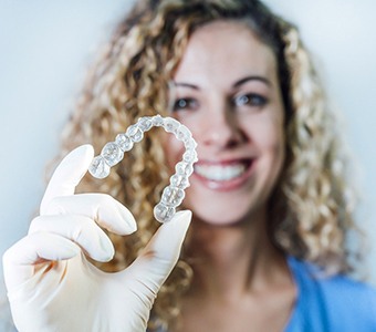 A woman with blonde, curly hair wearing a glove on her right hand and holding an Invisalign aligner