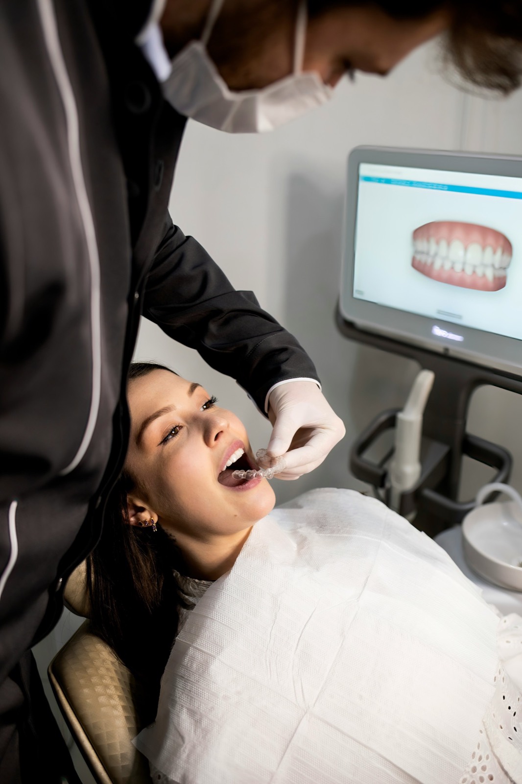 Dentist fitting a clear aligner while a 3D digital scan of the patient's teeth displays on the monitor